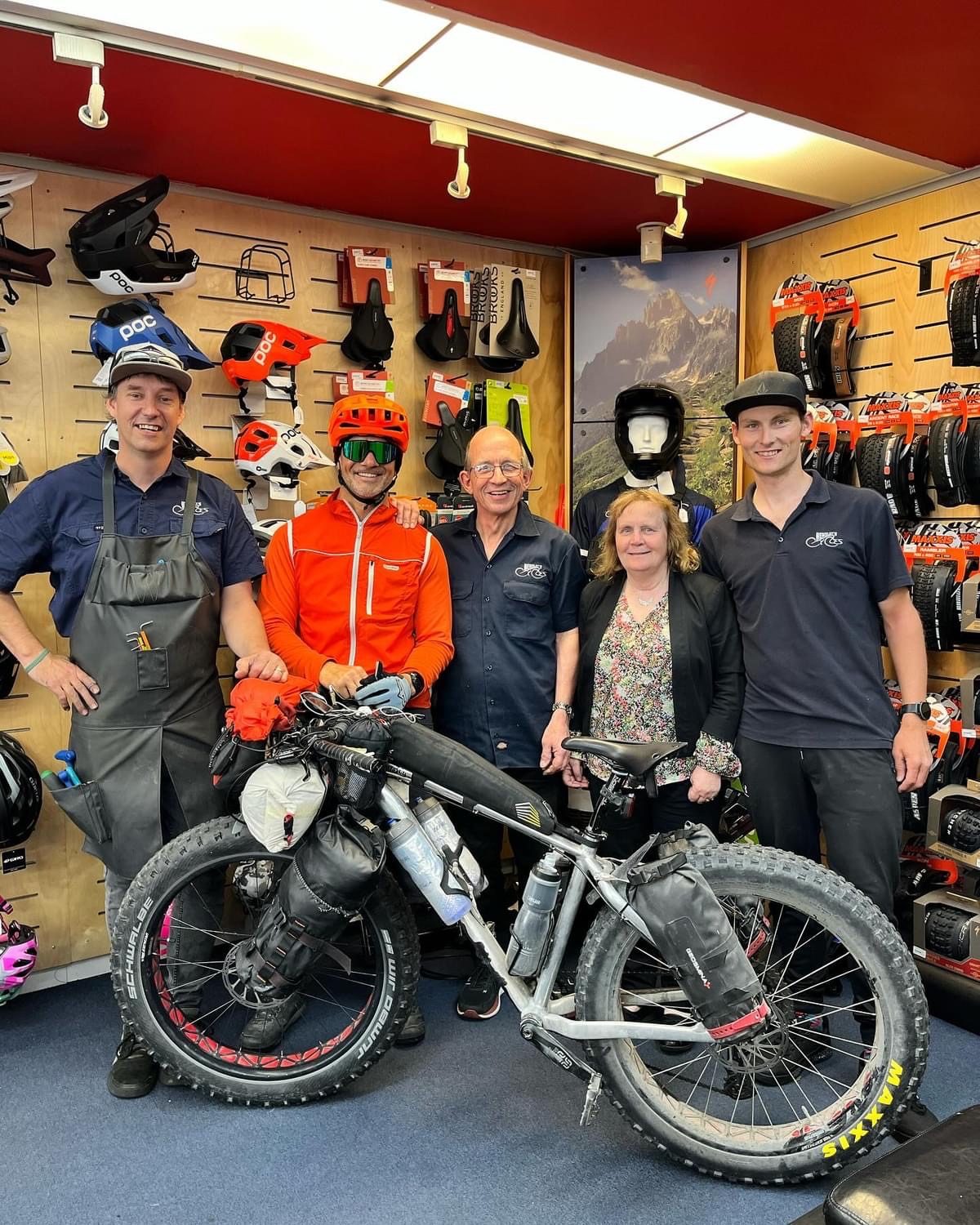 Doug in an orange jacket and helmet with his loaded fat bike, with the Wensleys Cycles team in their shop